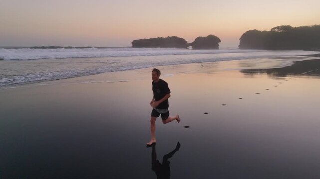 Drone view of a man running on a black sand beach at sunset with a sea arch in Bali. Cinematic active lifestyle and travel concept.