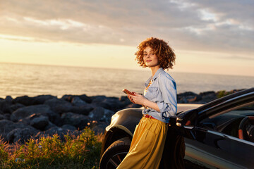 Fototapeta premium A young woman with curly hair stands beside a parked car by the rocky coast at sunset, exuding calm confidence and a relaxed, aspirational mood.