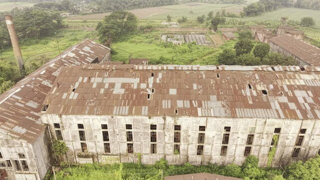Drone footage flying left to right across the abandoned Kushtia Sugar Mill, revealing damaged corrugated roofs, deteriorated factory buildings, and the inactive industrial complex.