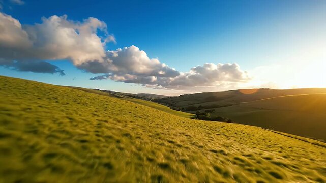 Vast sunlit grassy hillside under a clear blue sky with scattered fluffy clouds and distant rolling hills