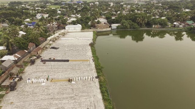 Forward drone footage documenting the Khajanagar rice processing area with drying yards, industrial buildings, access roads, and surrounding ponds in Kushtia, Bangladesh.