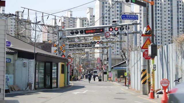 A street-level view of an urban Seoul road with railway crossing infrastructure, overhead signs, and pedestrians nearby.