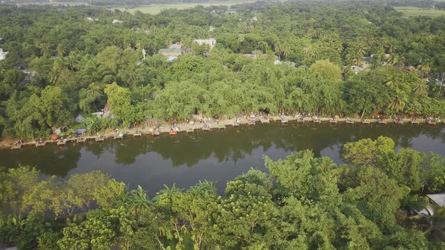 Aerial drone footage with a gentle right-to-left curve over a rural fishing competition in Bangladesh, revealing people along the pond bank, fishing stalls, calm water, and the landscape. 