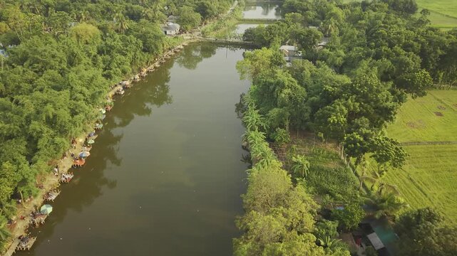 Aerial drone footage moving forward over a fishing competition at a rural pond in Bangladesh, showing people along the bank, fishing spots, calm water, and green countryside surroundings.
