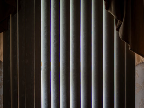 Slices of bright daylight peeking through closed vertical window blinds inside a dark room with a fabric curtain valance