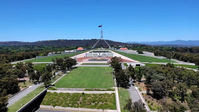 Aerial view of the iconic Parliament House with its distinctive flagpole, surrounded by lush green lawns and native trees in Canberra, Australian Capital Territory, Australia.