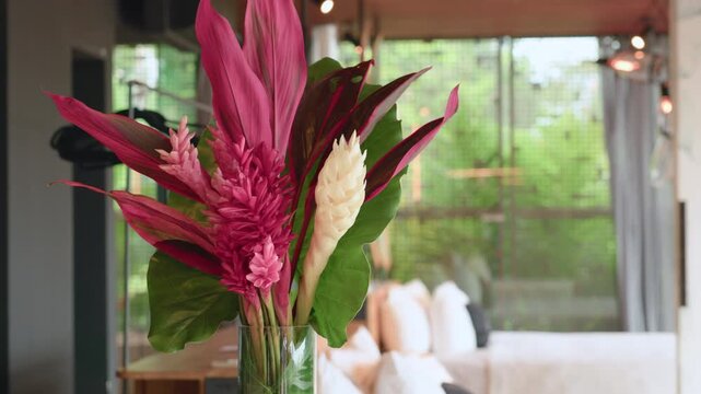 Vibrant tropical flower arrangement featuring pink and white ginger flowers with magenta ti leaves displayed in clear glass vase inside modern living space with natural light.