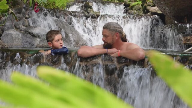 Father and young son share a peaceful moment relaxing together in a natural hot springs pool with tiered waterfalls cascading over volcanic rocks, framed by vibrant green tropical leaves in.