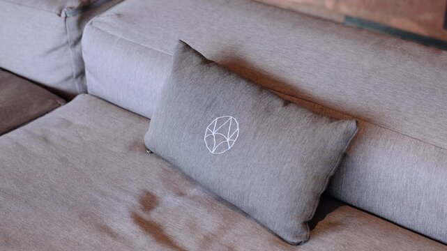 Close-up view of modern gray fabric sectional sofa with decorative throw pillow featuring white geometric embroidered pattern. Soft natural lighting highlights textile texture.