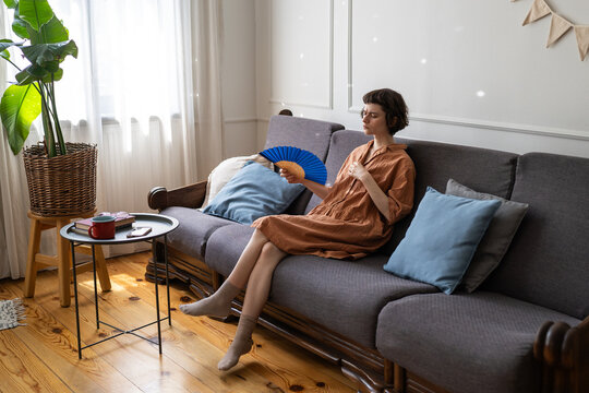 Unmotivated woman coping with summer heat in apartment with no air conditioning using hand fan for cooling, female with sick expression sits on couch staring and waving paper fan, hot season struggles