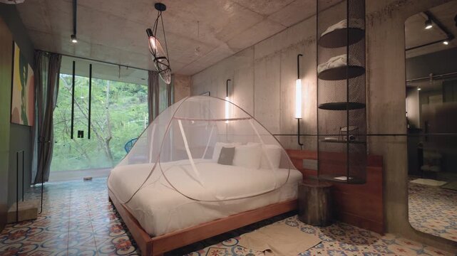 Stylish industrial loft bedroom featuring wooden bed with white mosquito net canopy and colorful patterned ceramic tile floor. Concrete walls with modern lighting and floor to ceiling windows.