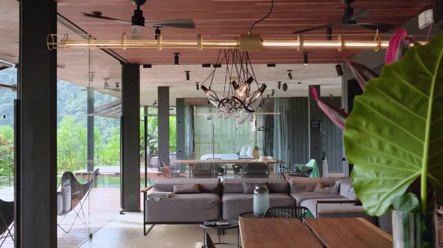 Luxurious open-plan living room in modern tropical villa featuring sectional sofa and designer pendant lights. Wooden ceiling with fans and glass walls reveal lush green jungle landscape outside.