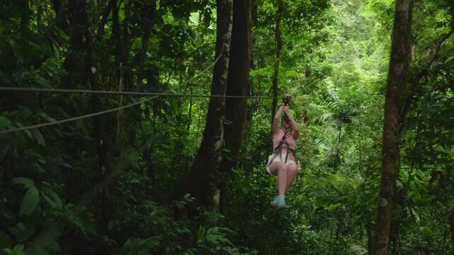 Girl on zipline soars through dense tropical rainforest wearing safety harness and green helmet.