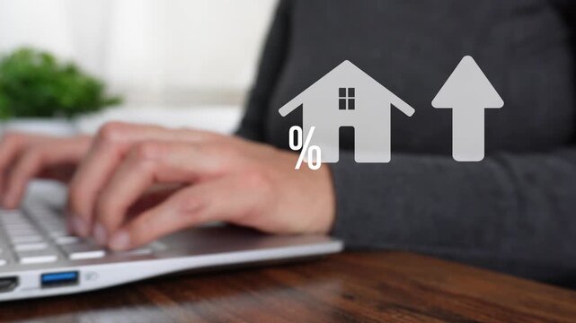 Close-up of a woman's hand working on a computer, pointing to an animated house icon with a percentage sign next to an upward-pointing arrow. Real estate theme.