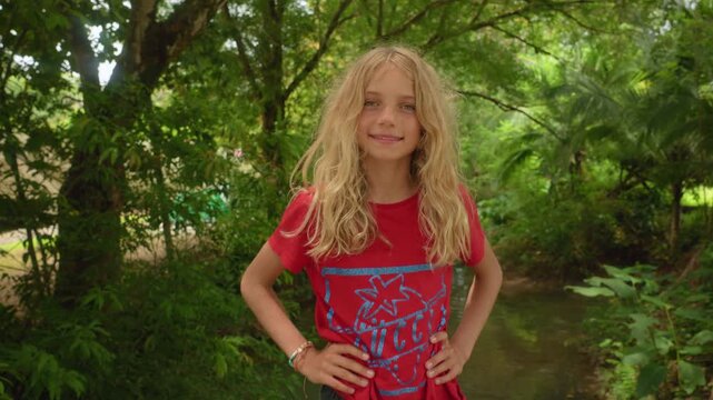 Young blonde girl with long wavy hair and green eyes smiles confidently at camera. She wears a red shirt and stands in a lush green garden with tropical foliage and stream.