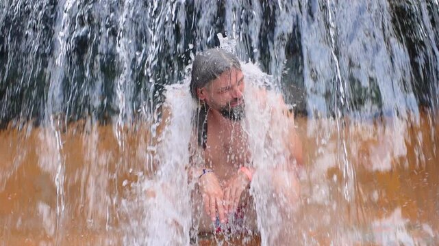 Slow motion footage of bearded man enjoying a refreshing waterfall experience as powerful cascading water splashes over his head and body against warm golden brown rock formations in tropical setting.