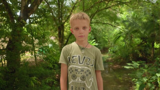 Portrait of a young blonde boy with green eyes wearing an olive t-shirt and pendant necklace standing in a lush tropical forest with stream and dense foliage in background.