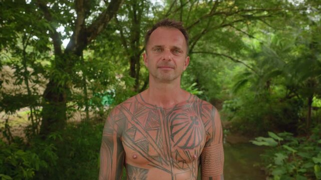 Middle-aged man with extensive geometric tribal tattoos covering his chest and arms stands shirtless in lush tropical jungle. Green foliage and trees create natural backdrop with soft daylight.