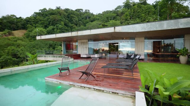 Stunning modern architecture villa featuring turquoise infinity pool with wooden deck and lounge chairs. Glass walls open to lush green tropical rainforest hills under overcast sky.