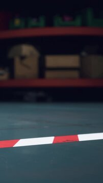 A worker sets down red and white safety markers on a warehouse floor. Boxes and shelves are visible in the background. The scene shows a busy work environment during daytime hours.
