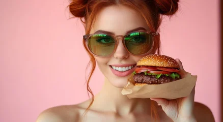 person with blurred face and red hair in space buns holding a sesame seed burger wrapped in paper against a pink background, playful indulgent mood © Rgrd