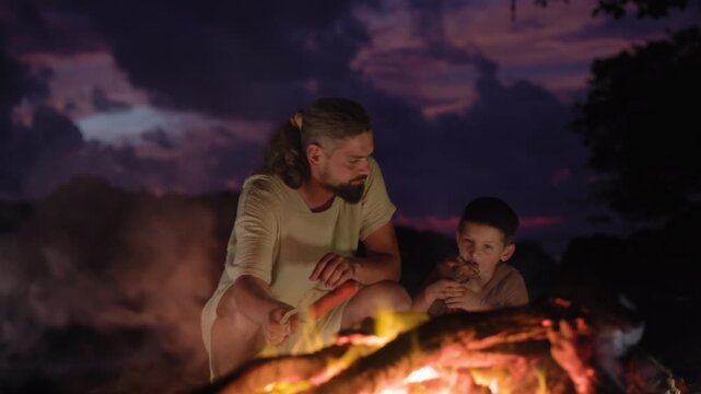 Father and son sit by a campfire on the ocean shore after sunset roasting sausages on wooden sticks. Warm flames illuminate their faces against dramatic purple twilight sky with glowing embers rising.