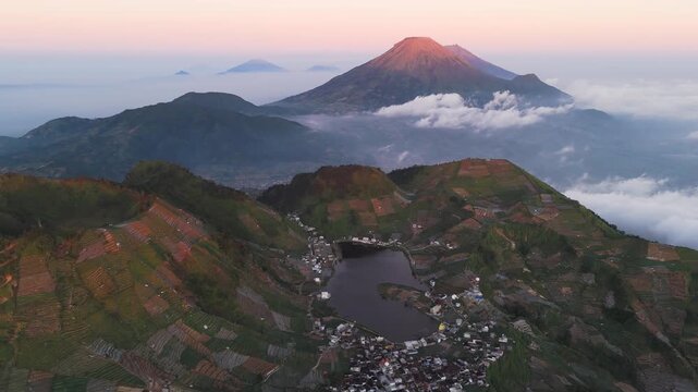 Aerial view of Telaga Cebong lake surrounded by terraced agricultural fields with the majestic Mount Sindoro volcano in the background at sunrise in Indonesia.