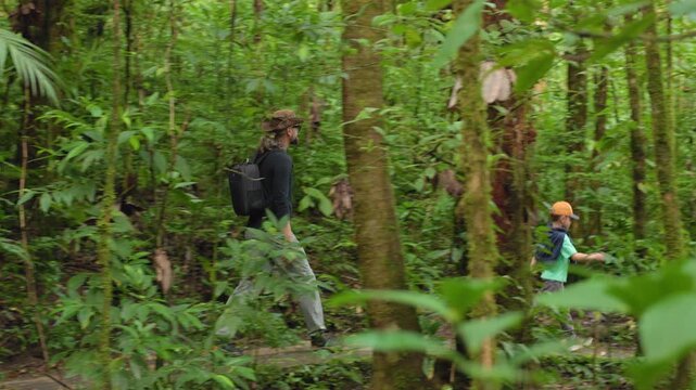 Father and son walk through jungle exploring lush tropical rainforest together. Dense green vegetation surrounds the pair as they hike along narrow trail with moss-covered trees and palm fronds.