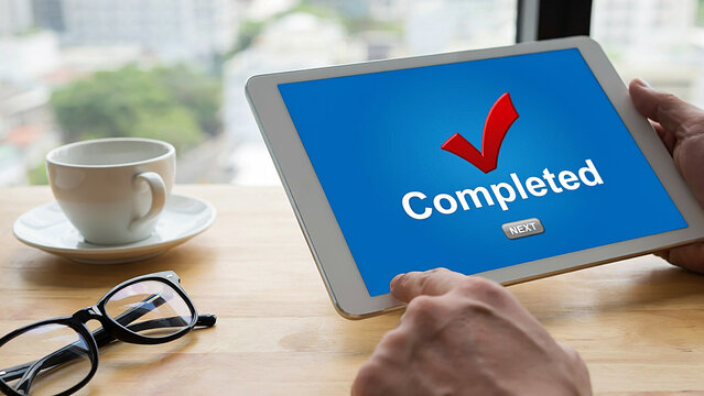 First person view of businessman holding tablet with completed checklist on screen on wooden desk with coffee cup