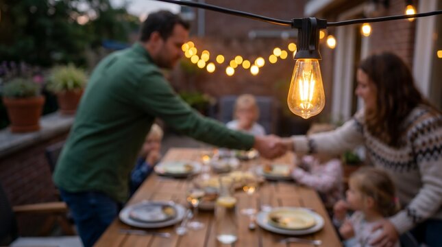 Family sharing a meal at outdoor wooden table under string lights at evening with father shaking hands with woman
