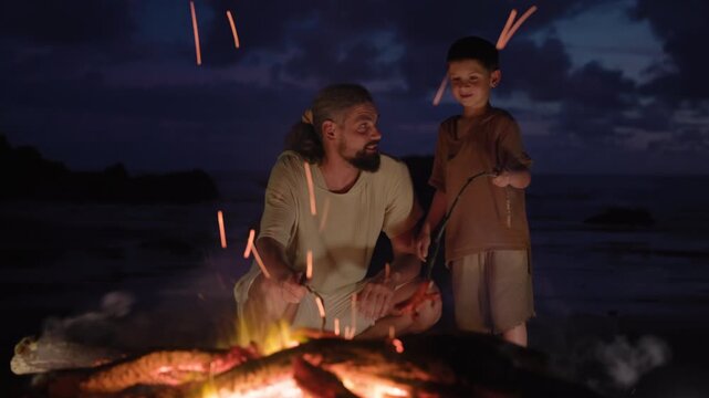 Father and son bond by a warm crackling campfire on a rocky ocean shore at twilight, roasting sausages on sticks while dramatic purple and blue clouds fill the dusky evening sky behind them.
