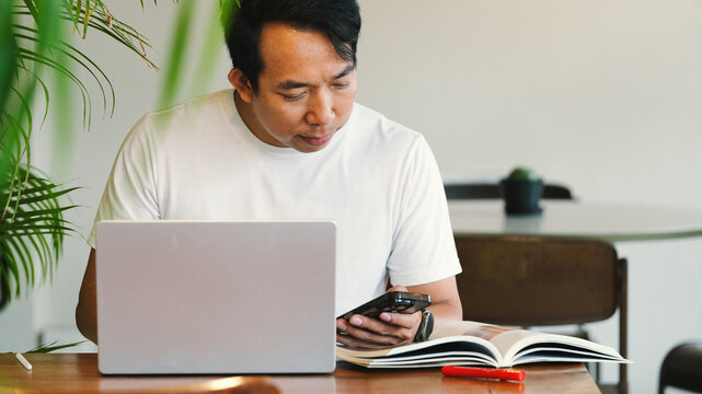 Focused Asian Businessman Using Smartphone and Laptop While Reviewing Notes at Modern Workspace