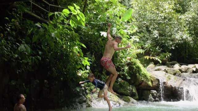 Father and son leap together from weathered metal staircase platform into emerald green natural swimming hole surrounded by dense tropical jungle foliage, moss-covered boulders.