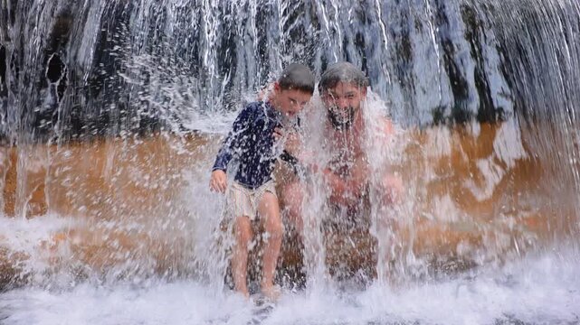 Father with son standing under powerful waterfall enjoying refreshing cascade in slow motion. Bearded dad holds young boy as water splashes around them creating joyful family bonding moment outdoors.