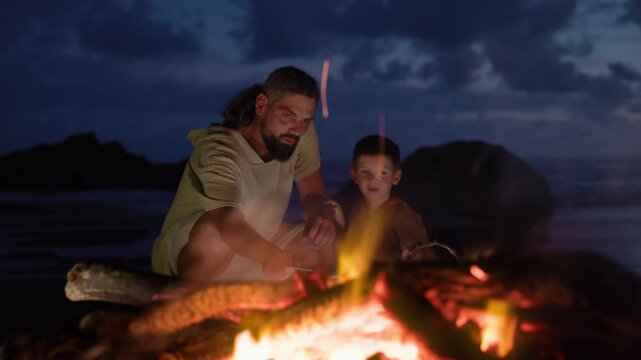 Father and son sit by campfire on ocean shore after sunset roasting sausages on sticks. Warm flames illuminate their faces against dramatic twilight sky with rocks and waves in the background.