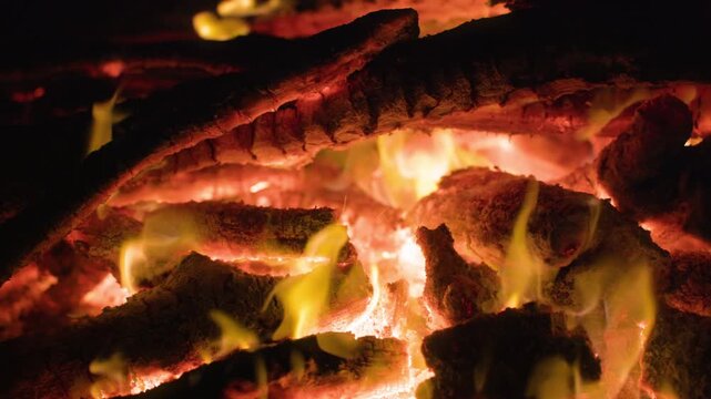Intense close-up footage of blazing firewood with bright yellow and orange flames flickering between charred logs. Glowing red-hot embers pulse with heat against a dark black background at night.