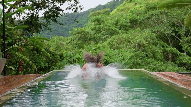 Slow motion footage of a man diving into an infinity pool surrounded by lush tropical rainforest and mountains. Water splashes dramatically as he emerges pushing wet hair back in scenic jungle.