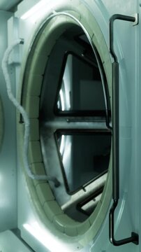 A washing machine door shows steam rising inside. The interior has metal parts and bright lights. A laundry room can be seen in the background with clear reflections on surfaces.