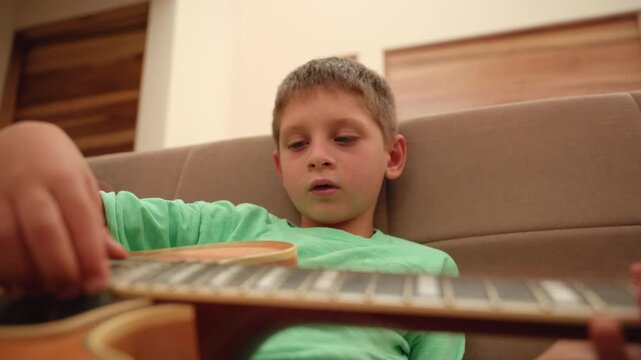 Young boy in green shirt practices acoustic guitar while relaxing on brown couch at home. Child focuses intently on fretboard and strings during music lesson in cozy living room setting.
