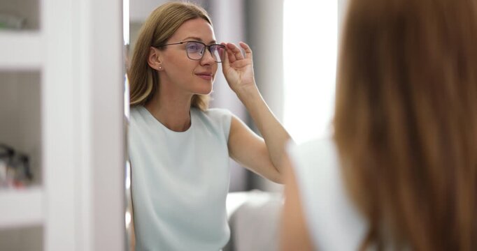 Young adult woman customer testing fashionable eyewear in optical boutique trying on selected frame smiling at mirror reflection checking fit comfort and personal style before making buying decision