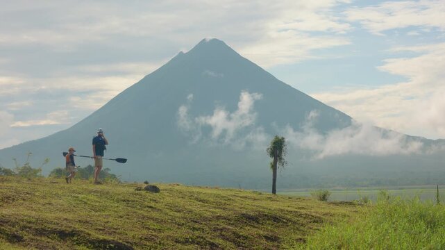 Father and son prepare for kayaking as they walk across grassy hillside carrying paddles with dramatic conical volcano rising in background surrounded by clouds and mist in tropical landscape.