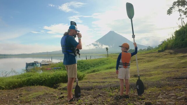 Father and son prepare for kayaking near a calm lake with a majestic volcano in the background. Both wear life jackets and hold paddles while standing on grassy shore during morning light.