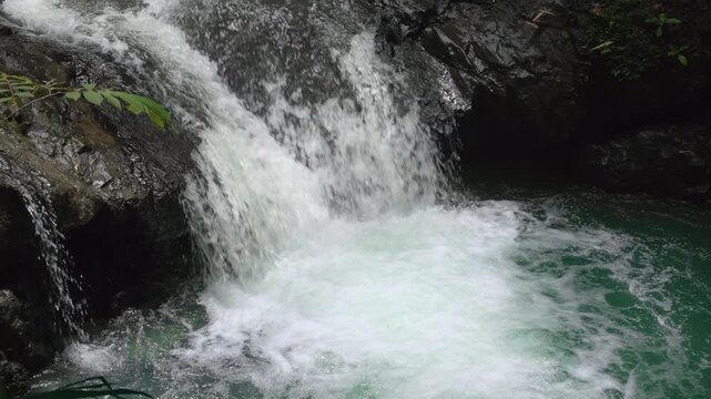 Powerful waterfall crashes over dark wet rocks into emerald turquoise pool creating white foam and spray. Lush green tropical foliage frames the cascading water in natural jungle setting.