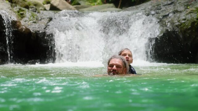 Father carries his young son on his back while swimming in a beautiful emerald green natural pool. A cascading waterfall splashes over mossy rocks in the lush tropical jungle setting.