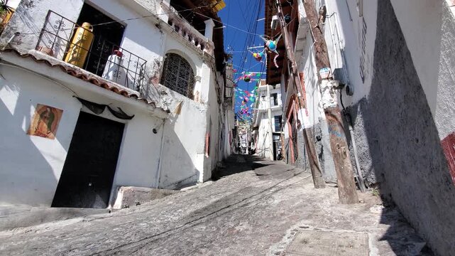 View of steep cobblestone street with white colonial buildings and hanging decorations in Taxco, Guerrero, Mexico.