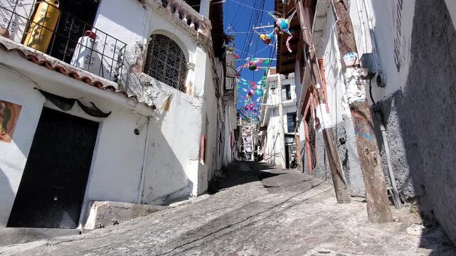 View of a narrow cobblestone street lined with white colonial buildings and colorful hanging decorations in Taxco, Guerrero, Mexico.