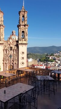 View of the ornate facade and twin bell towers of Santa Prisca de Taxco church, seen from a terrace with tables and chairs in Taxco, Guerrero, Mexico.