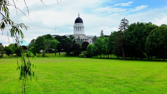 Maine State House capitol dome across a wide sunlit lawn framed by willow branches