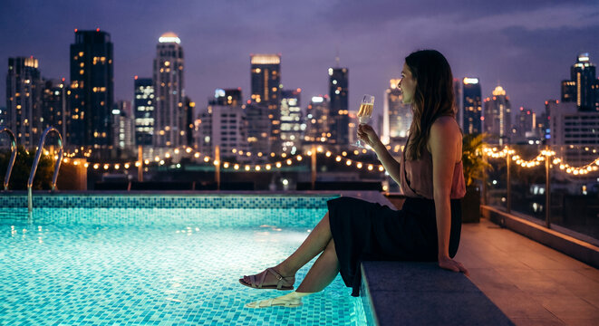 Woman with champagne by rooftop pool at night city view