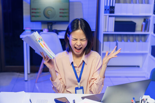 Stressed young Asian businesswoman overwhelmed with workload and holding documents while screaming in office.